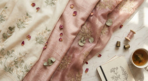 A flat lay of layered South Asian fabrics from cotton lawn to shimmering organza arranged on a marble surface with soft natural lighting.